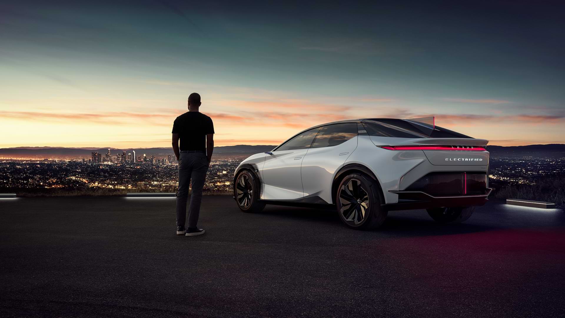 Man stands beside LF-Z on-top a hill, looking out across a city with the sun setting behind it.
