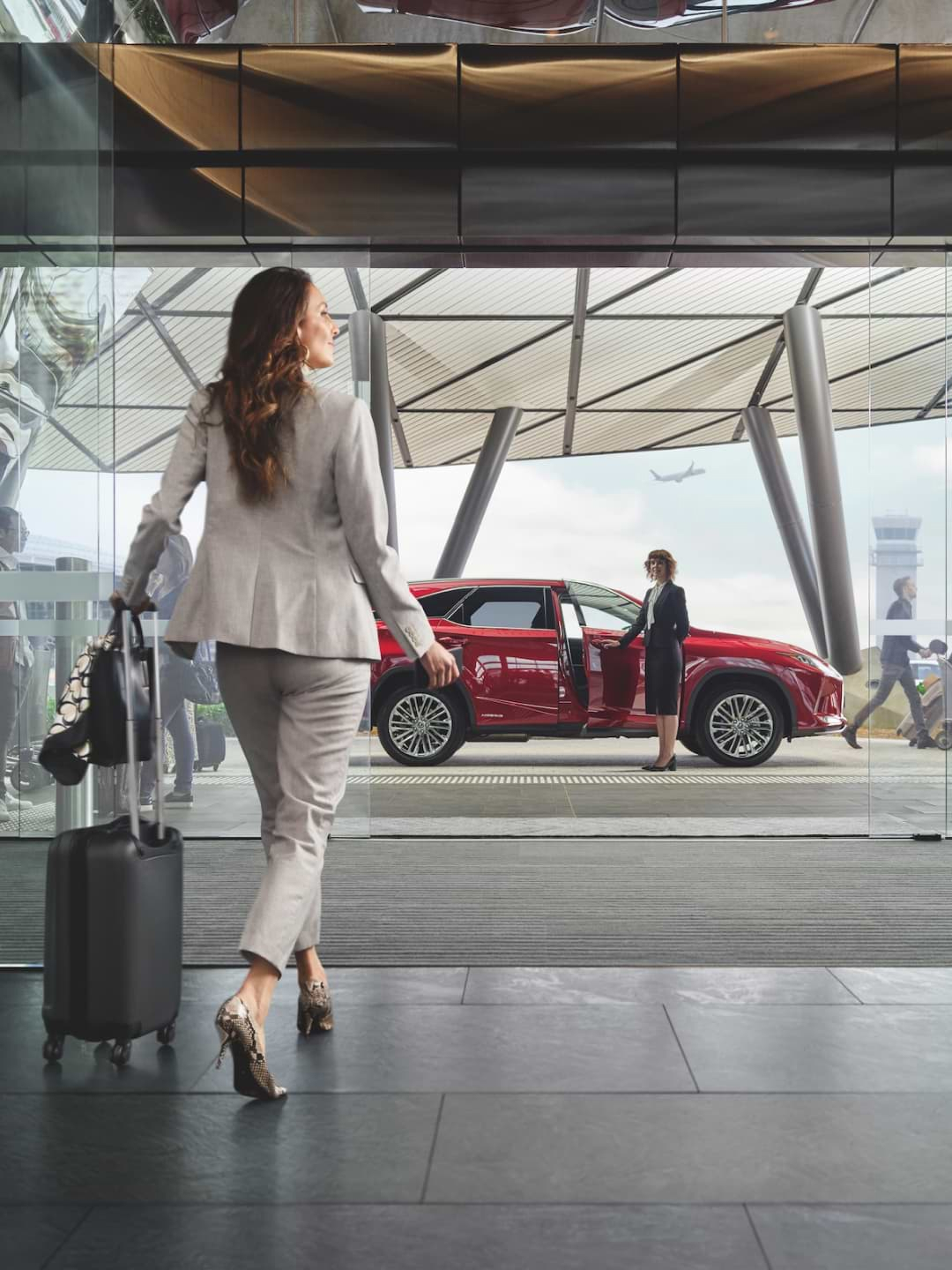 Woman wheels a suitcase out of an airport, as a red Lexus RX sits awaiting her for pickup.